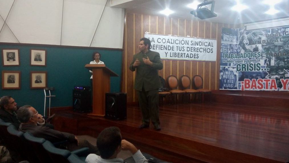 Frank Sandoval wearing all green and speaking emphatically on a raised platform indoors