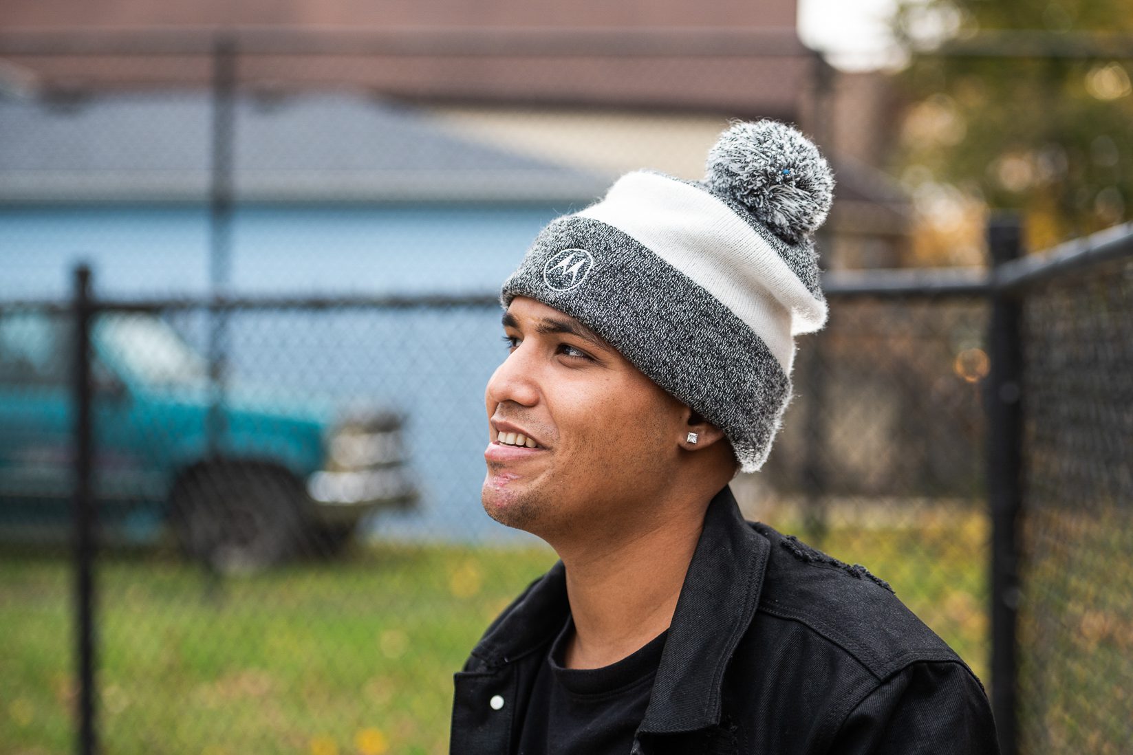 Nolram wears a gray and white beanie with a black shirt and jacket and black fence behind him
