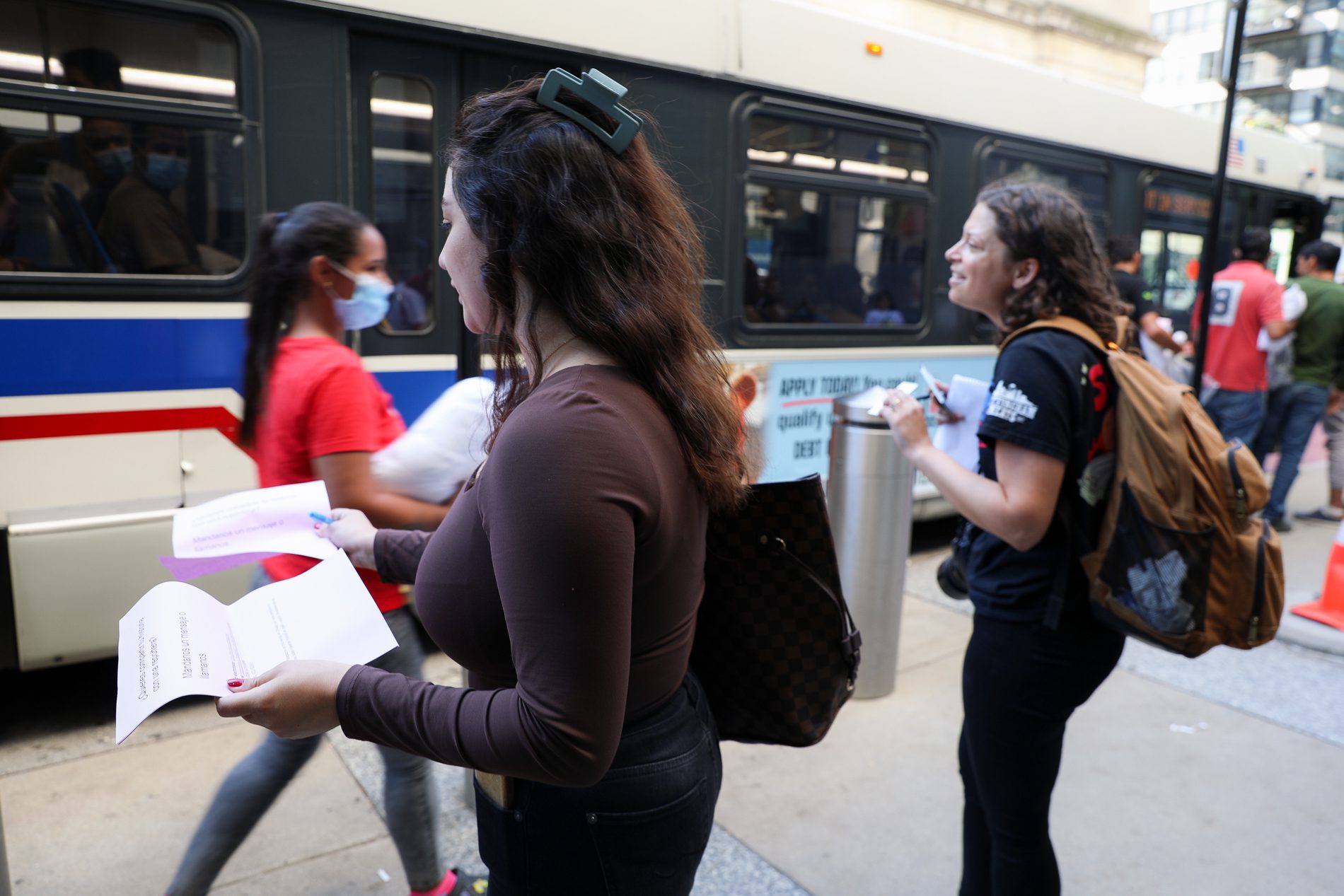 Diane wearing a brown long sleeve shirt hands out purple flyers to migrants walking from one bus to another; Michelle in black pants and a t-shirt with a tan backpack hands out business cards