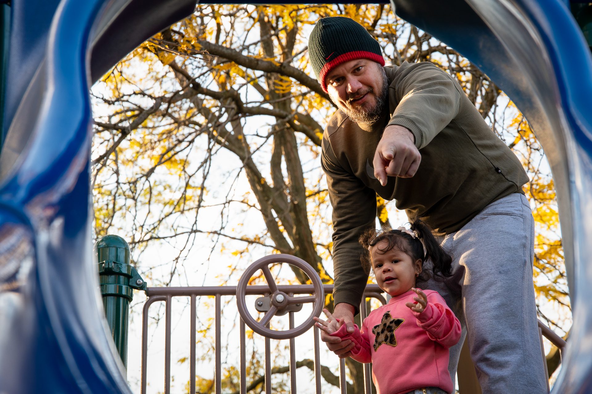 Tomas points Grecia towards a blue slide on the playground, she has a ping sweater with a star on it